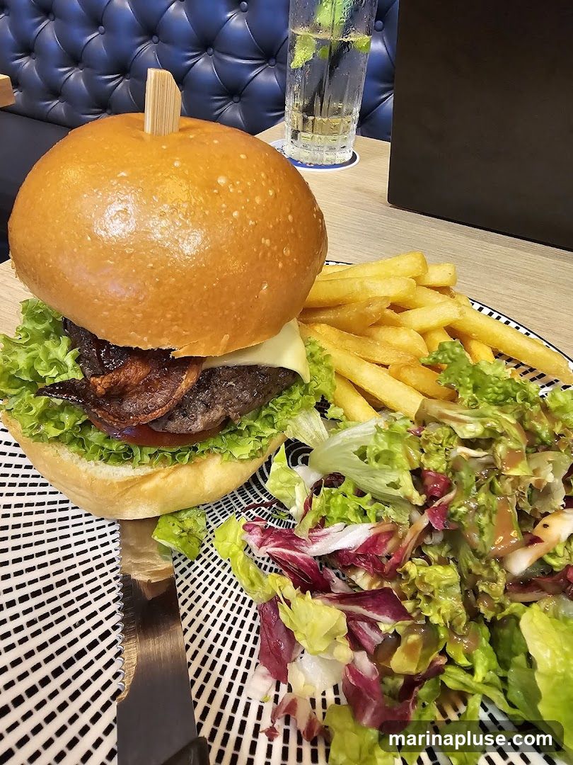 A close-up shot of a gourmet burger served with a side of french fries and a fresh salad. The burger has a golden-brown bun, lettuce, cheese, and what appears to be bacon. The fries are golden and crispy, and the salad is a mix of greens with a dressing. A glass with a drink and mint is visible in the background, along with a tufted blue seat.