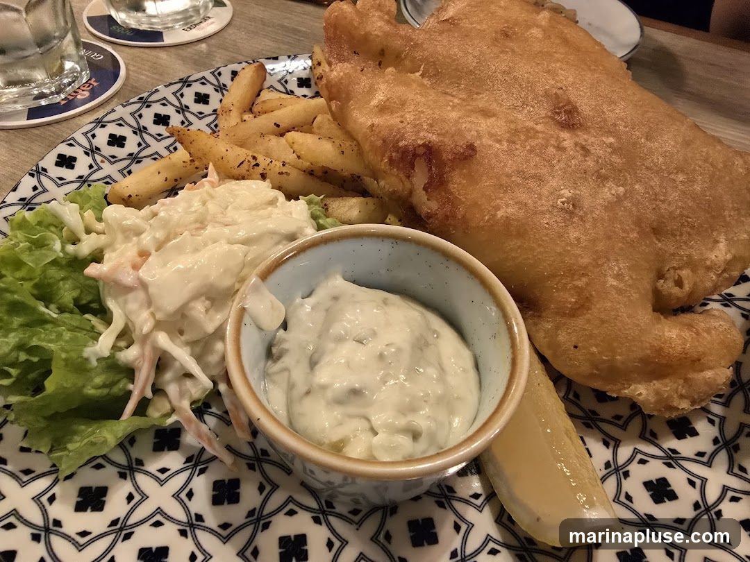 A close-up shot of a plate of fish and chips. The dish includes a large piece of battered fish, a side of french fries, coleslaw, a lemon wedge, and a small bowl of tartar sauce. The plate has a decorative blue and white pattern. In the background, there are coasters with logos, suggesting a pub or restaurant setting.