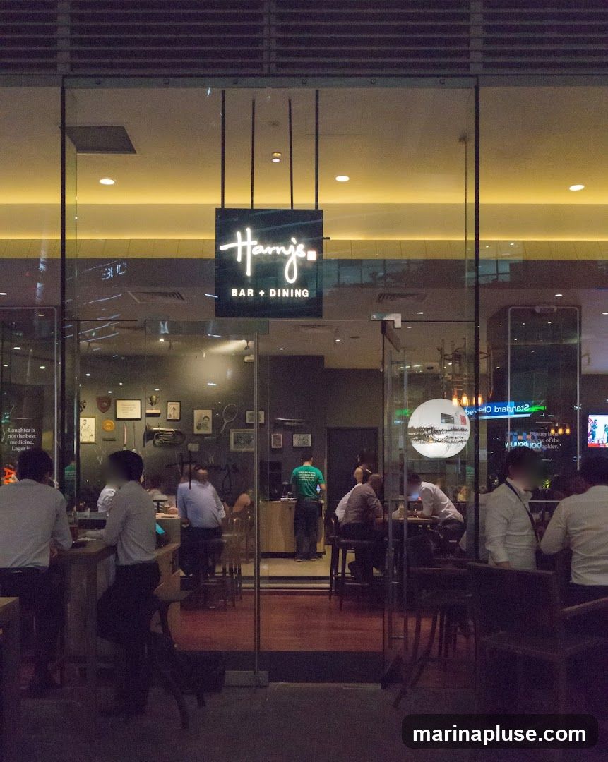 The exterior of Harry's Bar + Dining is visible at night. The entrance features large glass doors and windows, with a prominent black sign hanging above the entrance that reads "Harry's" in white script, followed by "BAR + DINING" in smaller white text. Inside, patrons are seated at tables and a bar, with some staff members visible. The interior has a warm ambiance with yellow lighting and decorative items on the walls.