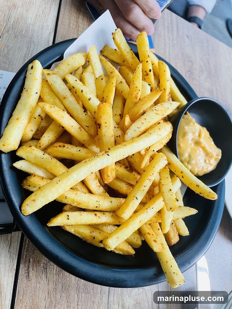 A close-up shot of a generous serving of seasoned french fries in a black bowl, accompanied by a small black bowl of dipping sauce. The fries appear golden brown and are sprinkled with visible herbs and spices. The dipping sauce is a creamy, light orange color. The image is taken from a top-down perspective, with a wooden table surface visible.