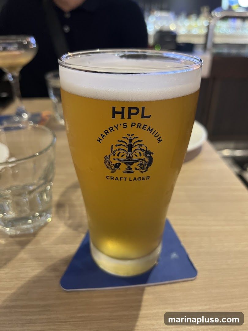A close-up shot of a tall glass filled with a golden-hued craft lager, topped with a frothy white head. The glass features a black logo with the text "HPL HARRY'S PREMIUM CRAFT LAGER" and an emblem of a fountain with mythical creatures. The glass sits on a blue coaster on a wooden table. In the background, out of focus, are other drinks and a person wearing a dark shirt.