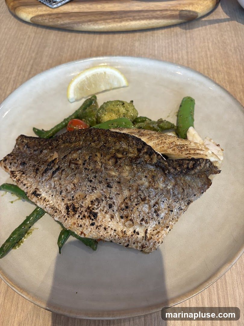 A close-up shot of a plate of grilled fish served with green beans and other vegetables, garnished with a lemon wedge. The fish has crispy skin and is seasoned with pepper. The vegetables appear to be blanched or lightly sautéed. The dish is presented on a light-colored, textured plate, with a wooden surface visible in the background.