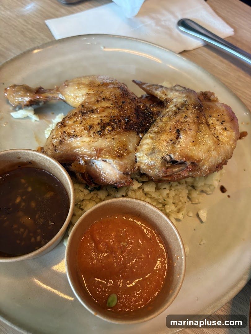 A plate with a half roasted chicken served over a bed of what appears to be rice or cauliflower rice. Two small paper cups contain dipping sauces, one dark brown and the other a reddish-orange. The chicken is golden brown with visible seasoning.