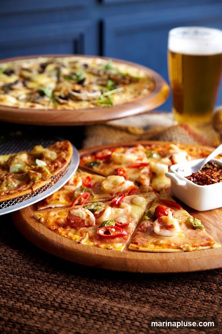 A close-up shot of several pizzas and a glass of beer. One pizza in the foreground is topped with shrimp, chili peppers, and herbs. Another pizza in the background appears to have mushrooms and greens. A small dish of red pepper flakes is also visible.