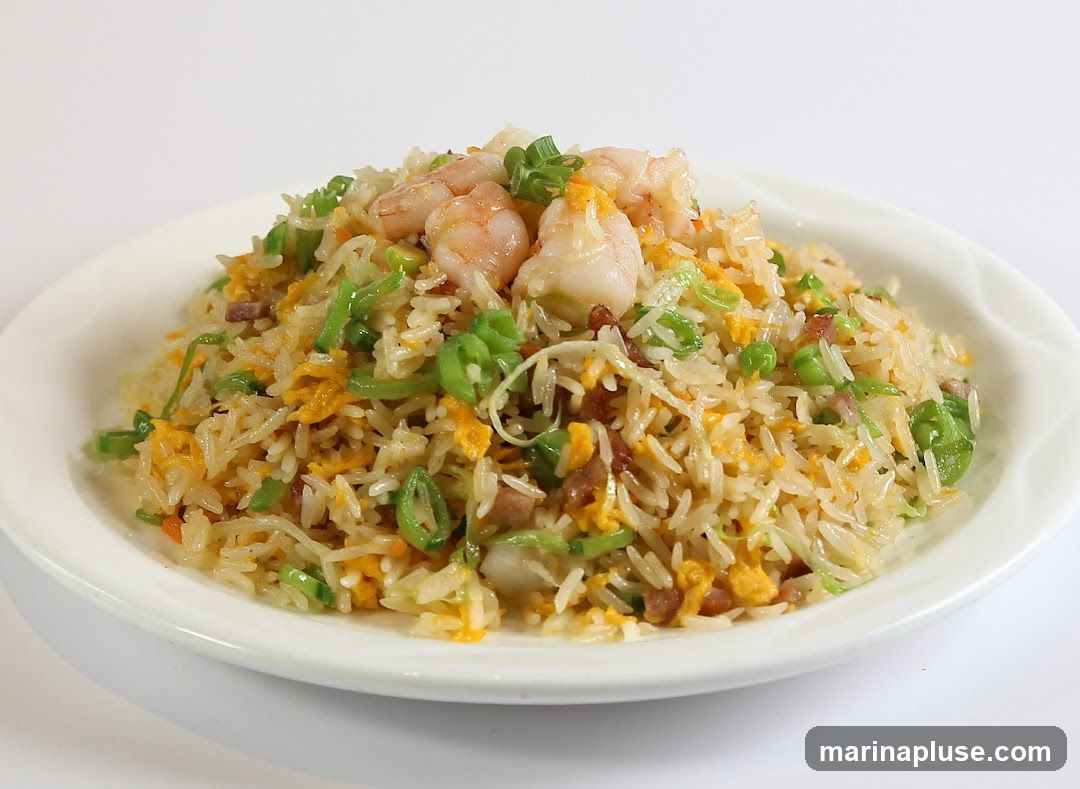 A white plate filled with shrimp fried rice. The rice is mixed with scrambled eggs, peas, chopped green onions, and small pieces of meat. Several cooked shrimp are arranged on top of the rice. The background is plain white.