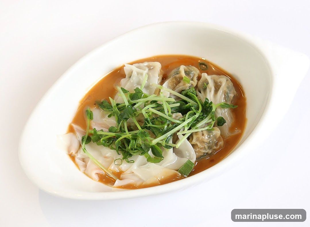 A close-up studio shot of wontons in a savory sauce, garnished with fresh sprouts and chopped scallions. The dish is served in a white, oval-shaped bowl against a plain white background.