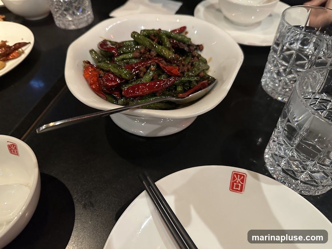 A close-up shot of a dish of spicy green beans with minced meat, served in a white bowl with a spoon. The dish is garnished with dried red chilies. In the foreground, there is a white plate with chopsticks and a small red emblem. Two glasses of water are visible on the right side of the image.