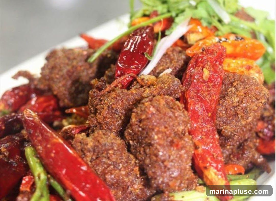 A close-up shot of a dish featuring crispy, fried beef pieces mixed with dried red chilies and green vegetables. The dish appears to be seasoned with spices, giving it a textured, reddish-brown appearance. Fresh green herbs are used as a garnish.