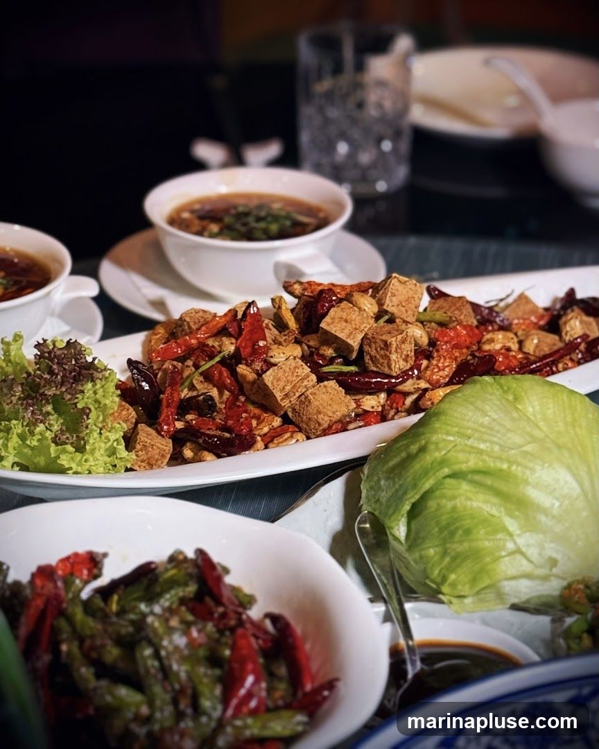 A close-up shot of several dishes on a table, likely in a restaurant. The main dish features cubed tofu stir-fried with dried chilies, nuts, and spices. To the left, a bowl of soup with green onions is visible, along with a small salad garnish. In the foreground, a bowl of green beans and red chilies is presented. A head of lettuce and a small dish of dipping sauce are also on the table. The setting appears to be a dimly lit dining environment.