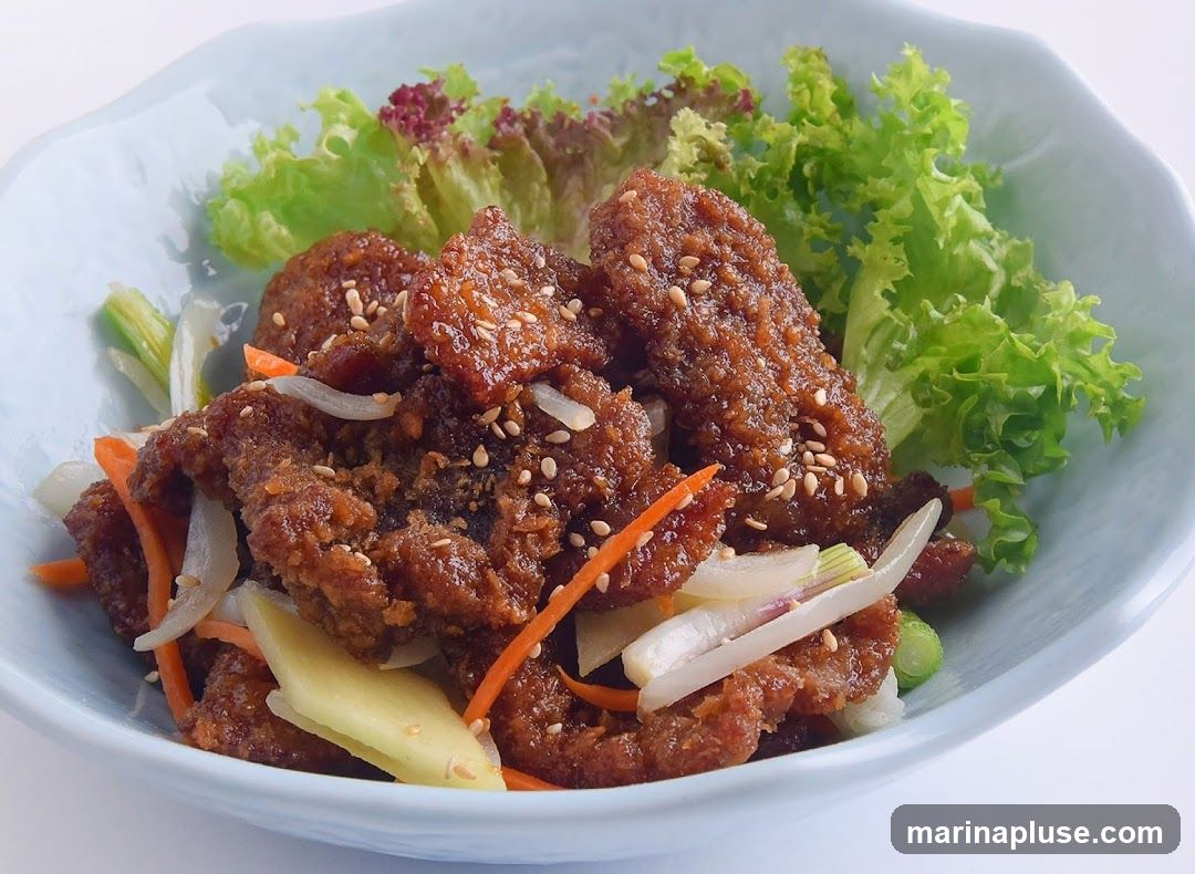 A close-up shot of a dish featuring crispy, glazed beef pieces sprinkled with sesame seeds. The beef is served with shredded carrots, sliced onions, and lettuce. The dish is presented in a light blue, wavy-edged bowl.