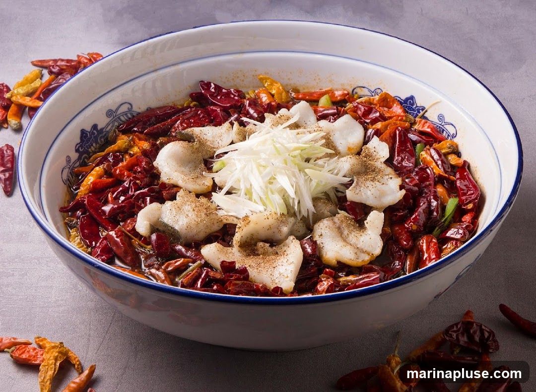 A close-up shot of a bowl filled with a spicy fish dish. The dish features white pieces of fish submerged in a generous amount of dried red chilies and other spices. Thinly sliced scallions are placed on top of the fish. The bowl has a blue and white decorative rim. Several dried chilies are scattered around the base of the bowl on a grey surface.