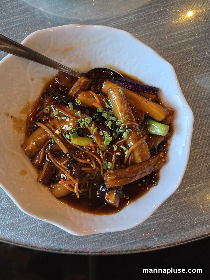 A close-up overhead shot of a white, wavy-edged bowl filled with braised eggplant pieces in a dark, glossy sauce. The dish is garnished with chopped green onions and julienned carrots. A spoon rests in the bowl. The table surface appears to be textured.