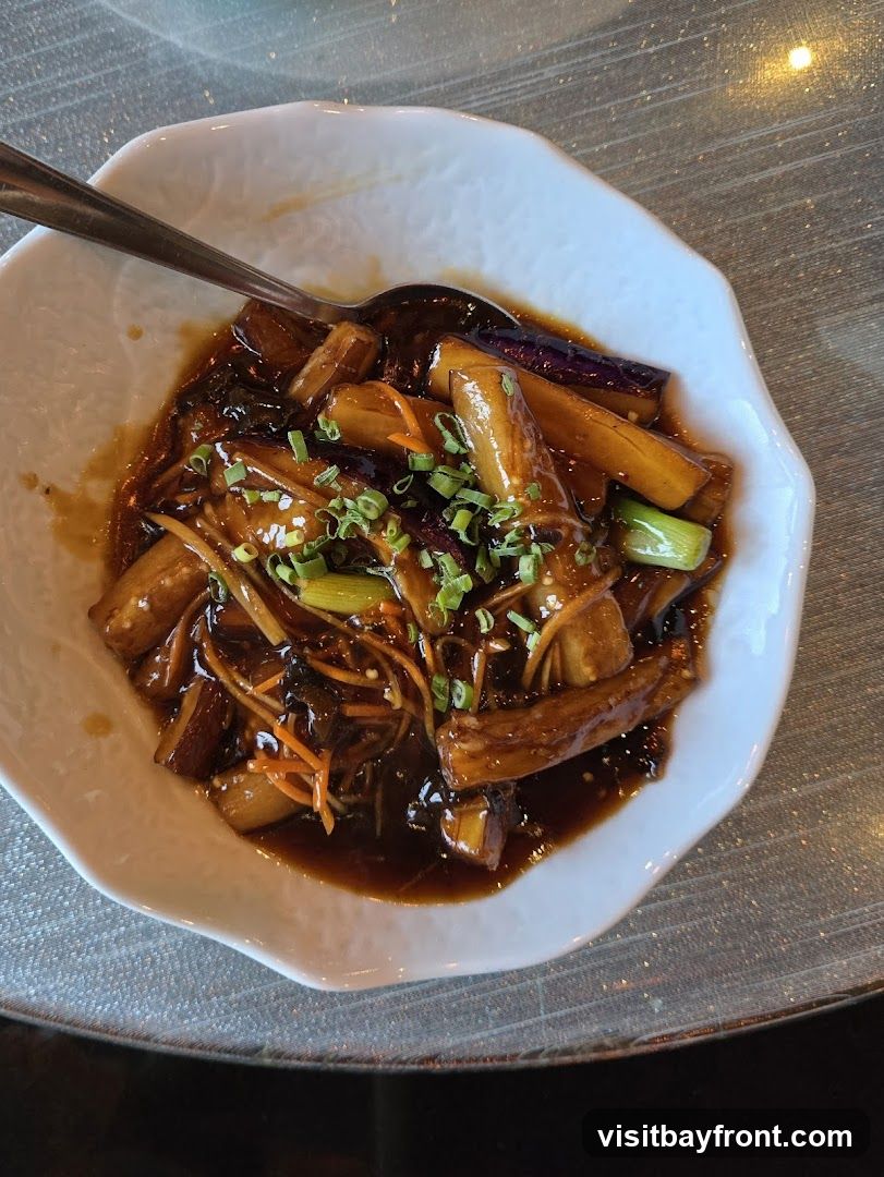 A close-up overhead shot of a white, wavy-edged bowl filled with braised eggplant pieces in a dark, glossy sauce. The dish is garnished with chopped green onions and julienned carrots. A spoon rests in the bowl. The table surface appears to be textured.