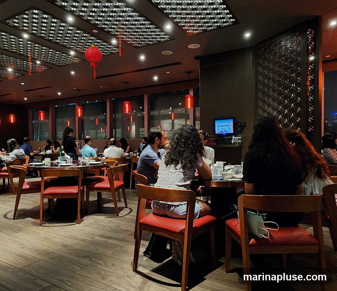 A dimly lit restaurant interior with several tables occupied by diners. Red lanterns hang from the ceiling, and decorative screens adorn the walls. A small screen is visible on a shelf in the background.