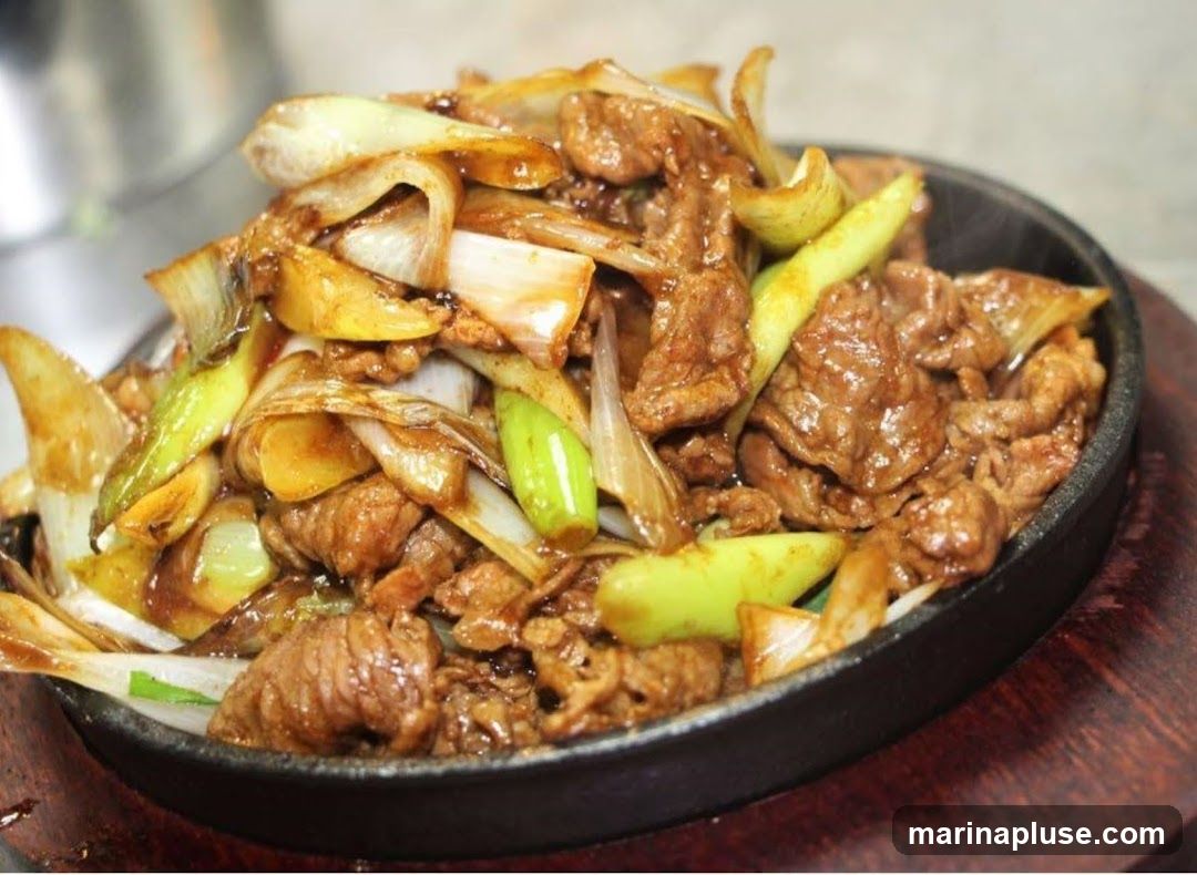 A close-up shot of a sizzling beef dish served in a cast-iron skillet. The dish features thinly sliced beef stir-fried with large pieces of white and green onions in a glossy brown sauce. The skillet is placed on a wooden base.