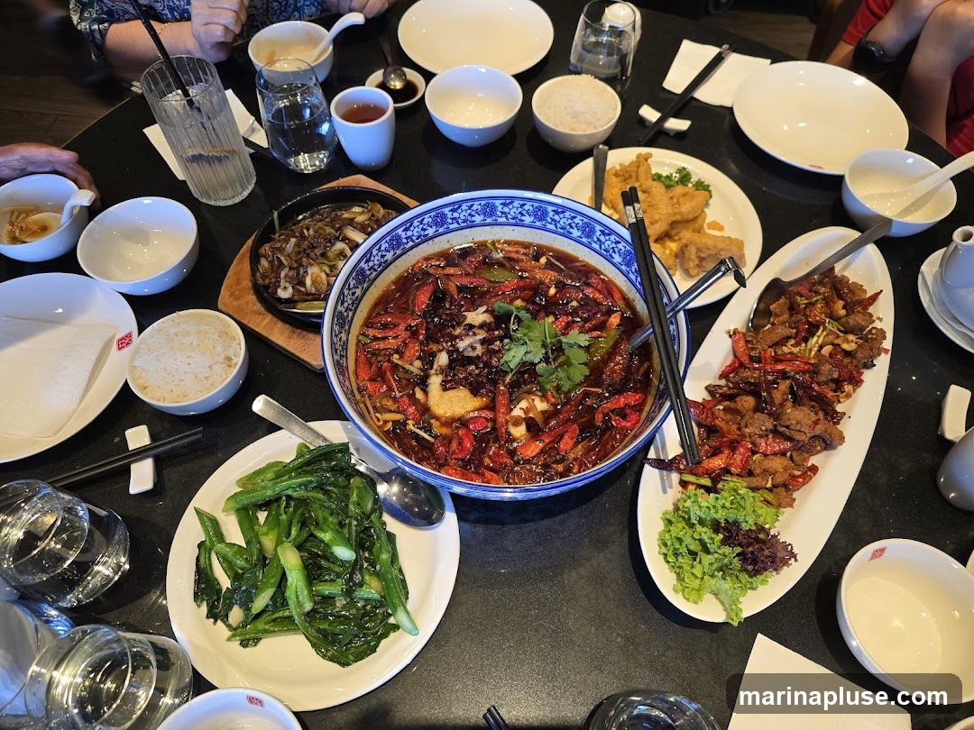 A table laden with various Chinese dishes, including a large bowl of spicy mala soup with chilies, a platter of crispy fried chicken, a dish of stir-fried vegetables, and a sizzling plate of meat and vegetables. Several bowls of rice, dipping sauces, and drinks are also visible, suggesting a shared meal.