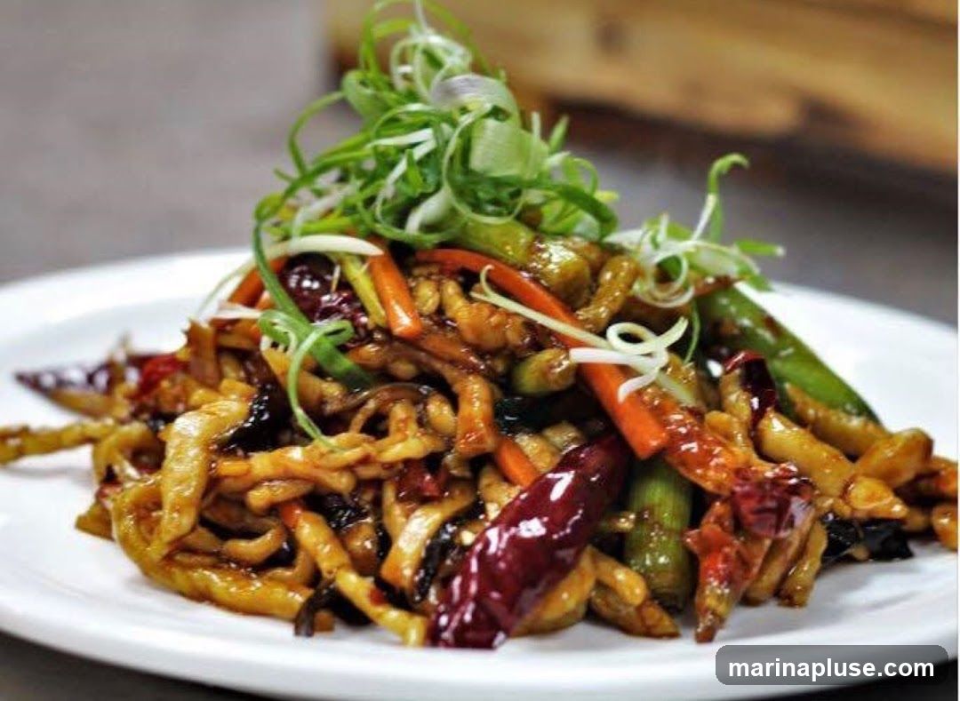 A close-up shot of a stir-fried dish served on a white plate. The dish appears to be made with shredded meat, julienned carrots, green beans, and dried chili peppers, all coated in a glossy sauce. It is garnished with thinly sliced green onions. The background is blurred, suggesting an indoor setting, possibly a restaurant.