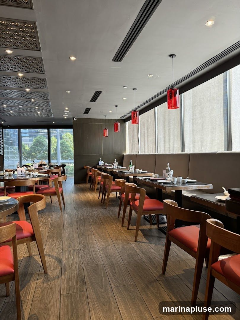 The interior of a restaurant with wooden floors and tables set for dining. Red chairs with wooden frames are arranged around the tables. Red cylindrical pendant lights hang from the ceiling. Large windows with blinds are visible along one wall, offering a view of trees and buildings outside. Decorative ceiling panels are also present.