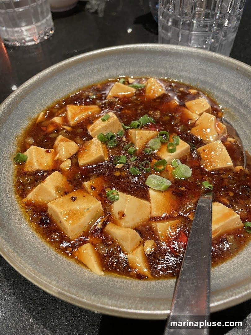 A close-up shot of a bowl of Mapo Tofu, a Sichuan dish. The tofu cubes are bathed in a rich, spicy sauce with visible chili flakes and green onions as garnish. A fork rests on the side of the bowl.