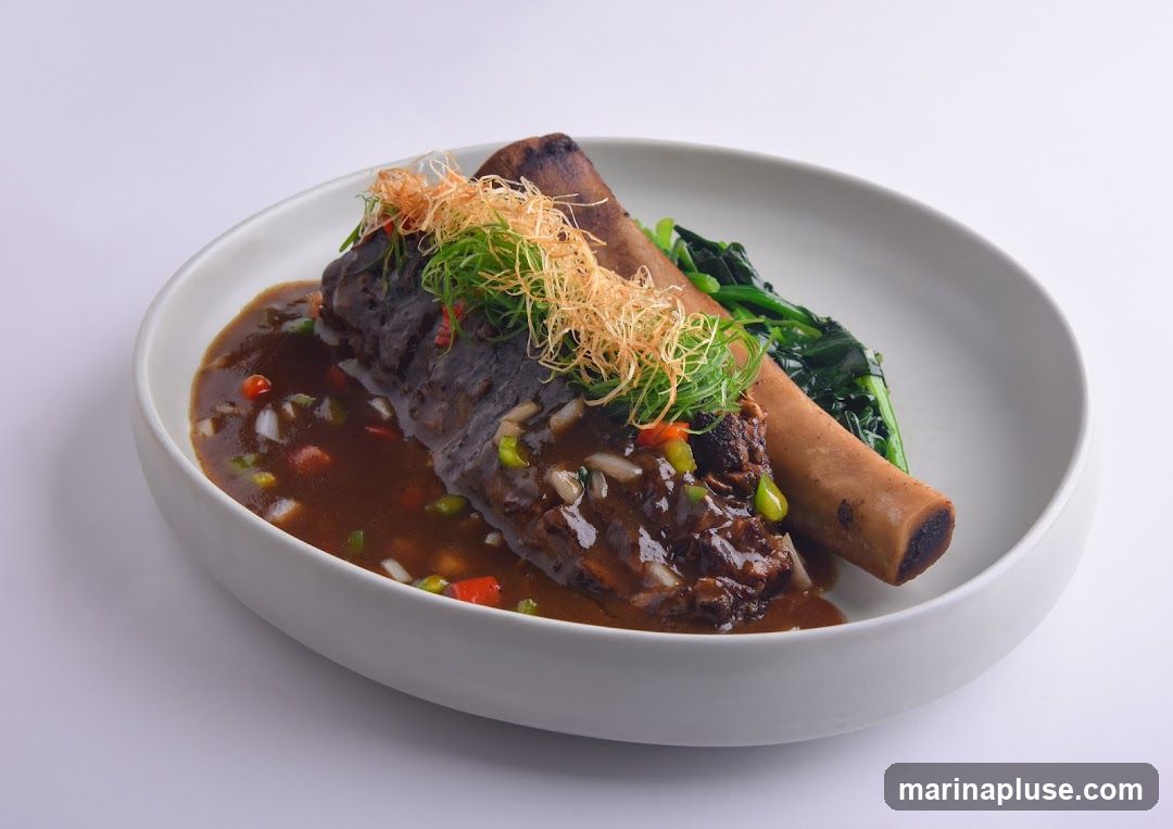 A close-up shot of a braised beef rib dish served in a white bowl. The beef rib is coated in a rich, dark sauce with visible pieces of onion, bell pepper, and chili. It is garnished with shredded fried onions and green onions. To the side, a portion of wilted green vegetables is visible. The dish is presented against a plain white background.