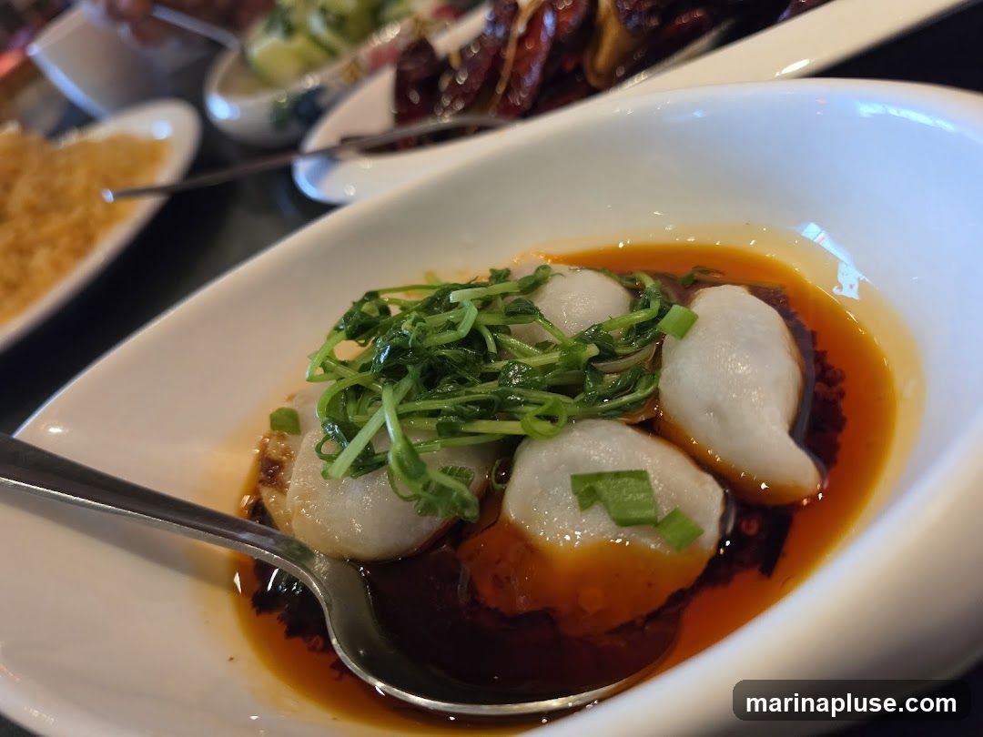 A close-up shot of fish balls served in a white bowl with chili oil and garnished with green sprouts and chopped scallions. A spoon is in the bowl. Other dishes are visible in the blurred background.