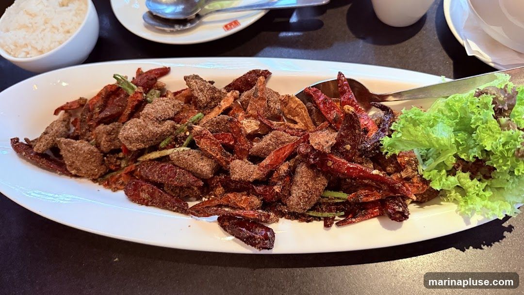 A white oval plate filled with a dish of fried ingredients, including many dried red chili peppers, and what appears to be seasoned meat or tofu. The dish is garnished with a small portion of lettuce on the right side. A bowl of white rice is visible in the upper left corner of the image. Spoons are also present on the table.