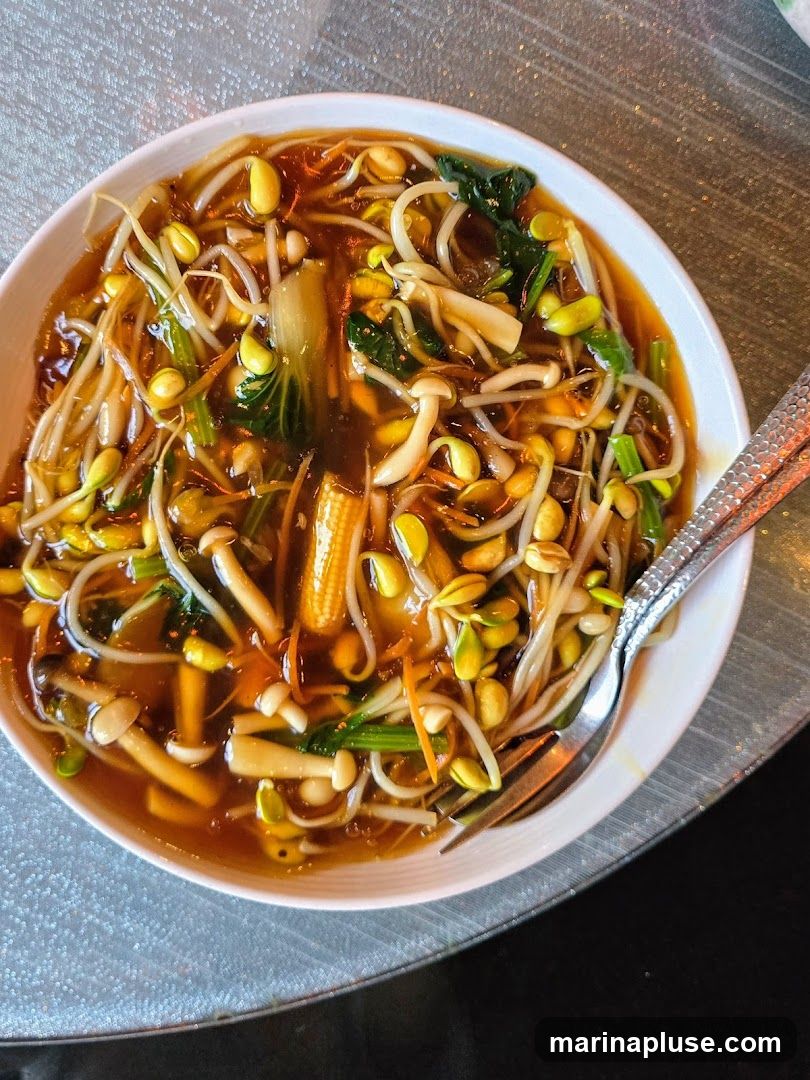A close-up overhead shot of a white bowl filled with a stir-fry dish. The dish contains a rich brown sauce, bean sprouts, bok choy, enoki mushrooms, shredded carrots, and small pieces of corn. A fork and spoon are resting on the side of the bowl. The table surface appears to be textured and metallic.