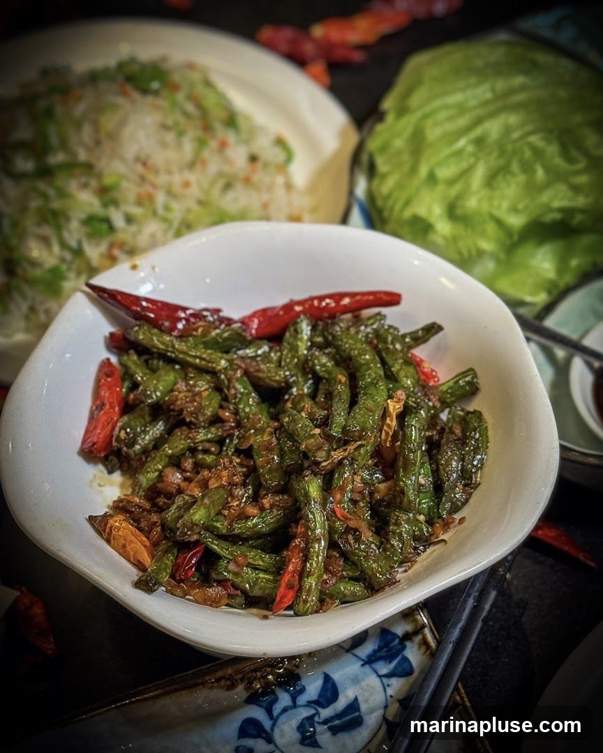 A close-up shot of a white bowl filled with stir-fried green beans, mixed with minced garlic and dried chili peppers. The beans are glossy and appear to be seasoned with a savory sauce. In the background, a plate of white rice with green vegetables and a head of lettuce are visible, along with scattered dried chili peppers.