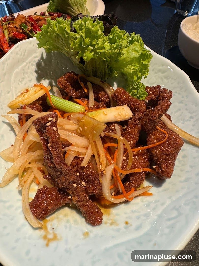 A close-up shot of a plate of crispy beef stir-fried with onions, carrots, and scallions, garnished with sesame seeds and served with lettuce leaves. Another dish with dried chilies and vegetables is visible in the background, along with a bowl of white rice.