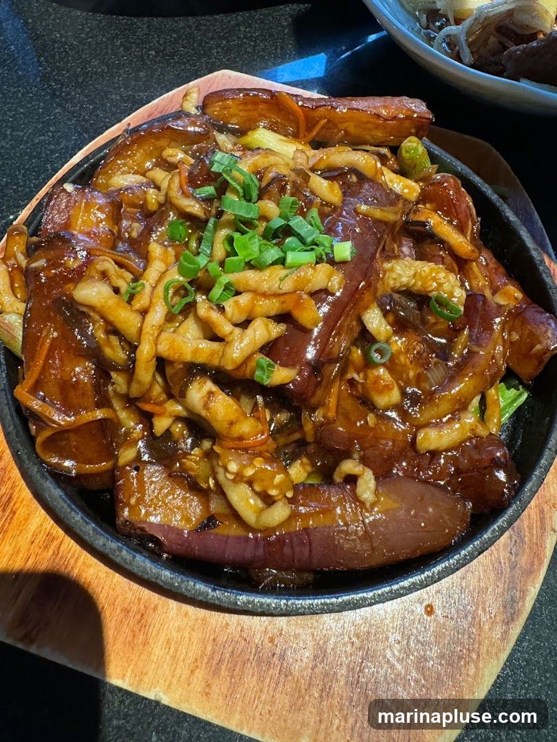 A close-up shot of a sizzling eggplant dish served in a black cast-iron skillet on a wooden board. The dish features thick slices of eggplant coated in a glossy, dark sauce, mixed with shredded vegetables and topped with chopped green onions. Another dish with noodles and meat is visible in the blurred background.