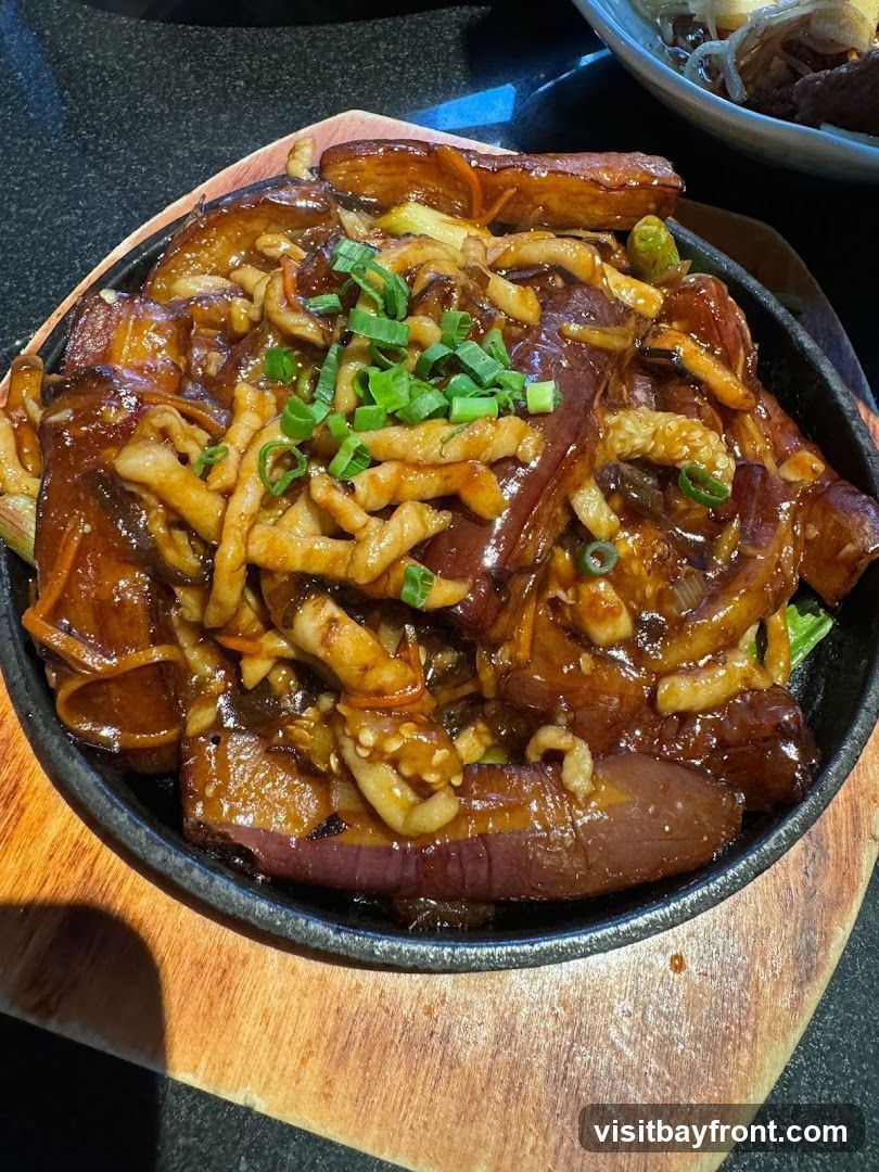A close-up shot of a sizzling eggplant dish served in a black cast-iron skillet on a wooden board. The dish features thick slices of eggplant coated in a glossy, dark sauce, mixed with shredded vegetables and topped with chopped green onions. Another dish with noodles and meat is visible in the blurred background.