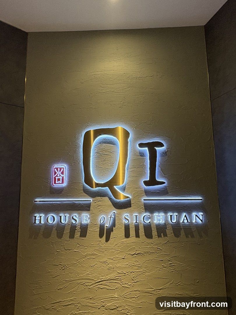 A close-up shot of a lit sign that reads "Qi HOUSE of SICHUAN". The sign is mounted on a textured wall.