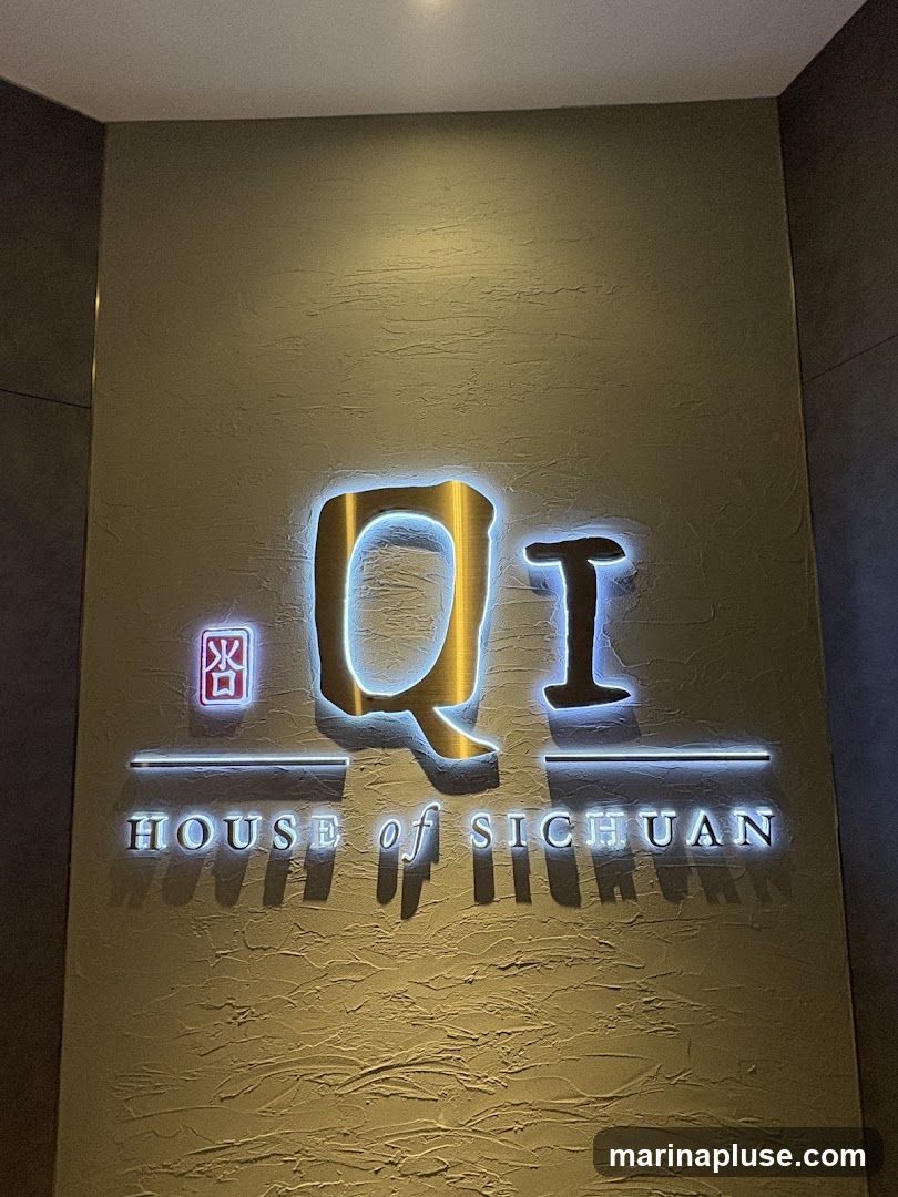 A close-up shot of a lit sign that reads "Qi HOUSE of SICHUAN". The sign is mounted on a textured wall.