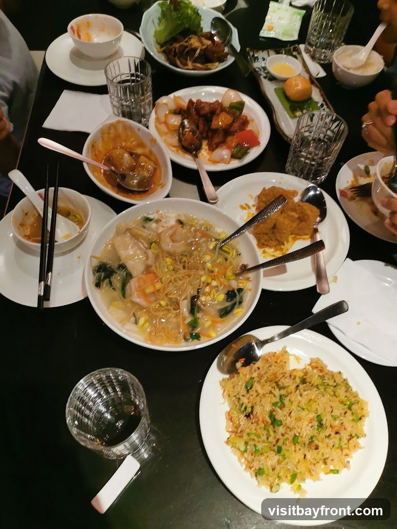 A top-down view of a table laden with various Asian dishes, including fried rice, noodles with seafood, and several saucy stir-fries. Several glasses and used bowls suggest a meal in progress.