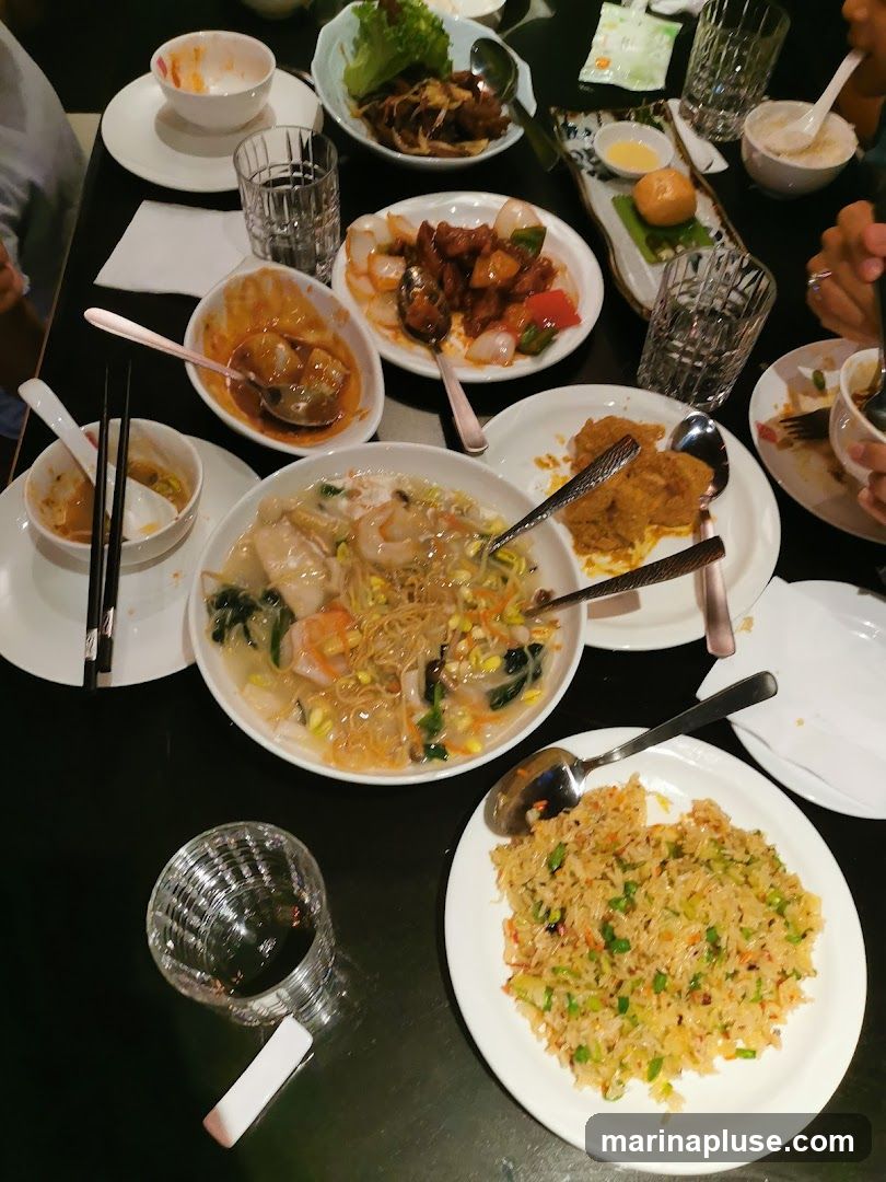 A top-down view of a table laden with various Asian dishes, including fried rice, noodles with seafood, and several saucy stir-fries. Several glasses and used bowls suggest a meal in progress.