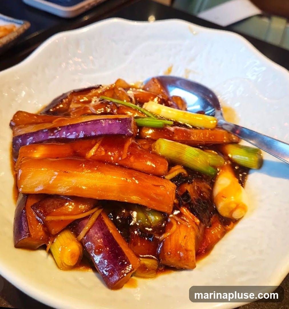 A close-up shot of braised eggplant and scallions in a glossy sauce, served in a white, scalloped-edge bowl. A spoon rests in the dish.