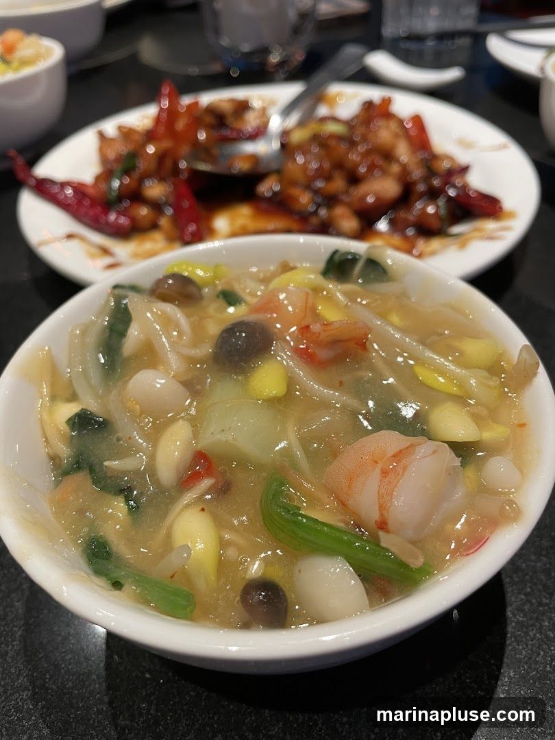 A close-up shot of two dishes of Chinese food. In the foreground is a white bowl filled with a light-colored soup containing shrimp, mushrooms, and vegetables. In the background, a white plate holds a dish of stir-fried meat with chili peppers and scallions.