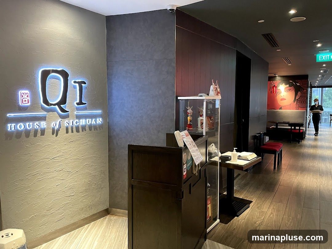The image shows the interior of a restaurant with a prominent sign that reads "Qi HOUSE OF SICHUAN". The sign is illuminated and features a stylized logo. To the right of the sign, there is a reception desk with a glass partition, hand sanitizer, and some decorative items. Further down the hallway, there are tables and chairs, and a large mural of a woman's face. An employee is visible walking down the hallway towards the exit.