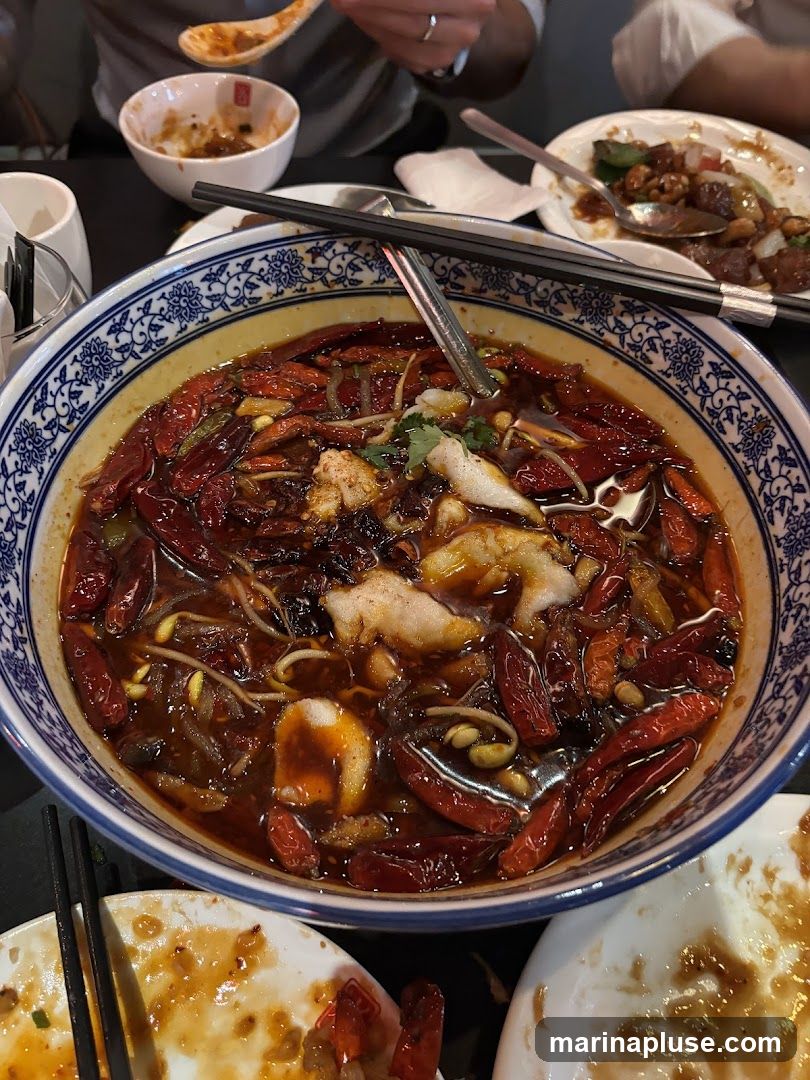 A close-up shot of a large bowl filled with a spicy fish dish. The dish contains chunks of white fish, bean sprouts, and numerous dried red chili peppers in a dark, oily broth. Chopsticks are resting on the rim of the bowl. Several plates with remnants of food are visible around the main dish, suggesting a shared meal.