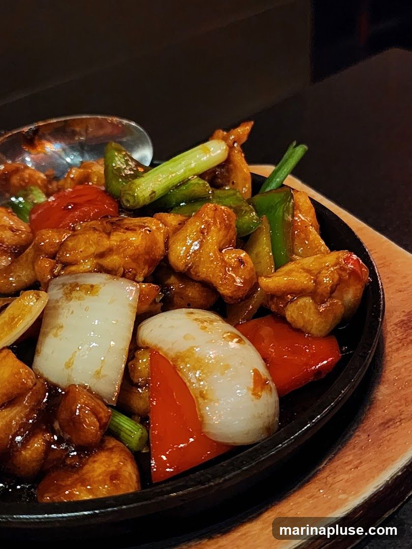 A close-up shot of a sizzling chicken dish served in a black cast-iron skillet on a wooden board. The dish features pieces of glazed chicken, red bell peppers, onions, and green onions. A spoon is visible in the upper left corner.
