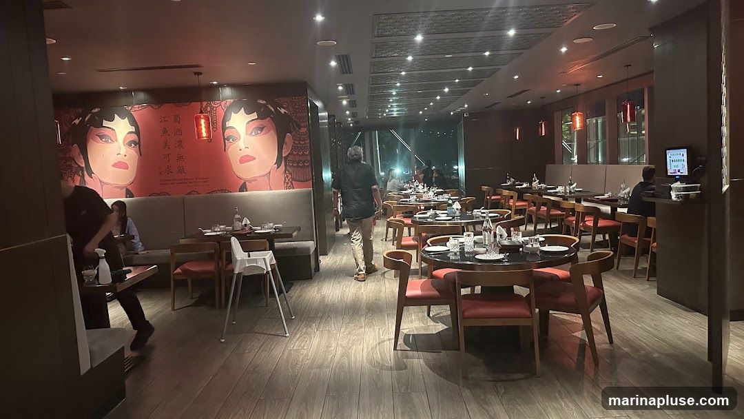 The image shows the interior of a restaurant with a modern design and Asian-inspired decor. The walls are adorned with large, stylized portraits of Peking Opera performers. The dining area features numerous tables set for meals, with chairs upholstered in red. Lighting fixtures hang from the ceiling, and the floor is made of wood-look tiles. Several patrons are visible, as well as staff members attending to tables. A digital menu or ordering screen is mounted on a pillar in the foreground.