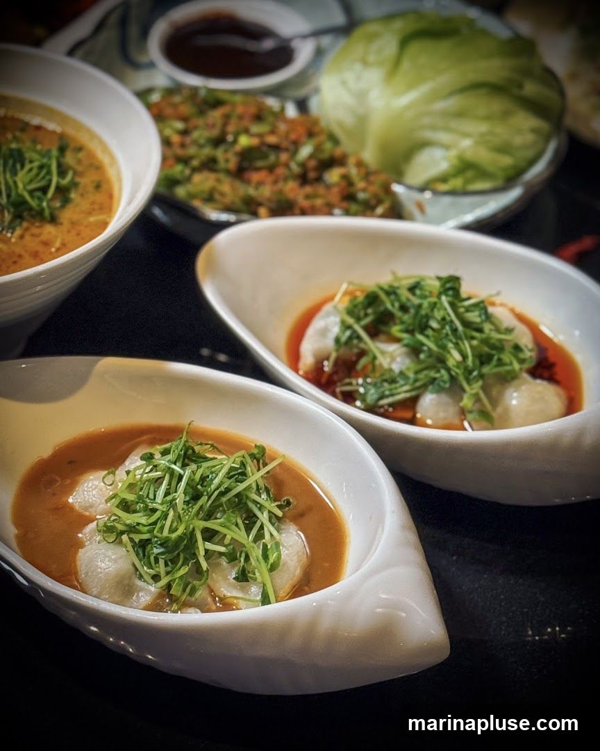 A close-up shot of several dishes of Asian cuisine. In the foreground, two white, boat-shaped bowls contain dumplings in a savory sauce, topped with fresh green sprouts. Behind them, a larger bowl holds a creamy, orange soup, also garnished with sprouts. Further back, a plate of what appears to be stir-fried vegetables and a head of lettuce are visible, along with a small dish of dipping sauce. The food is artfully presented on a dark surface.