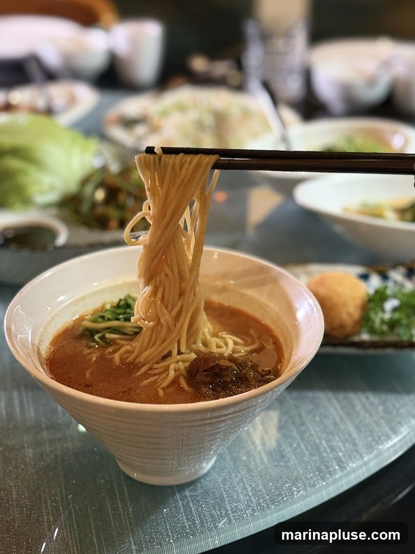 A close-up shot of a bowl of noodles being lifted with chopsticks. The noodles are thin and appear to be in a savory broth with some green garnishes and meat. In the background, other dishes are visible on a table, suggesting a meal setting.