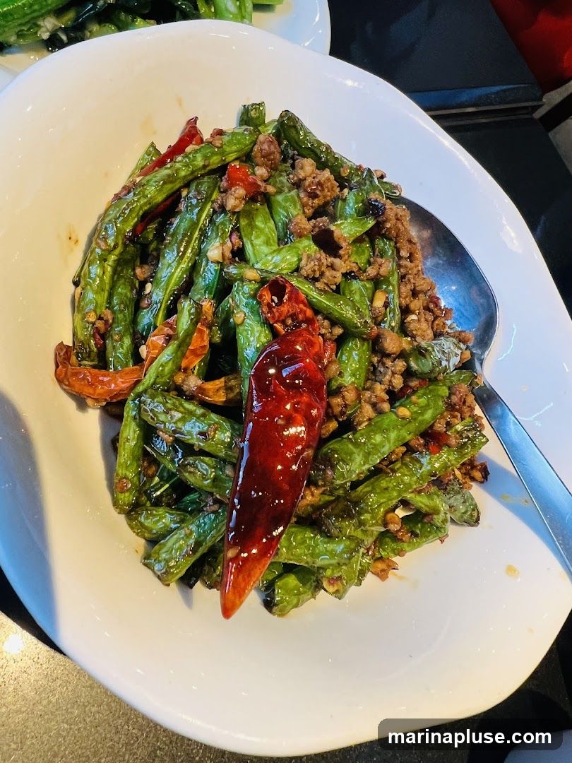 A close-up shot of a white bowl filled with stir-fried green beans, minced meat, and dried chili peppers. A large spoon rests in the bowl, ready to serve. The dish appears to be seasoned and slightly oily, with a glossy sheen on the beans.