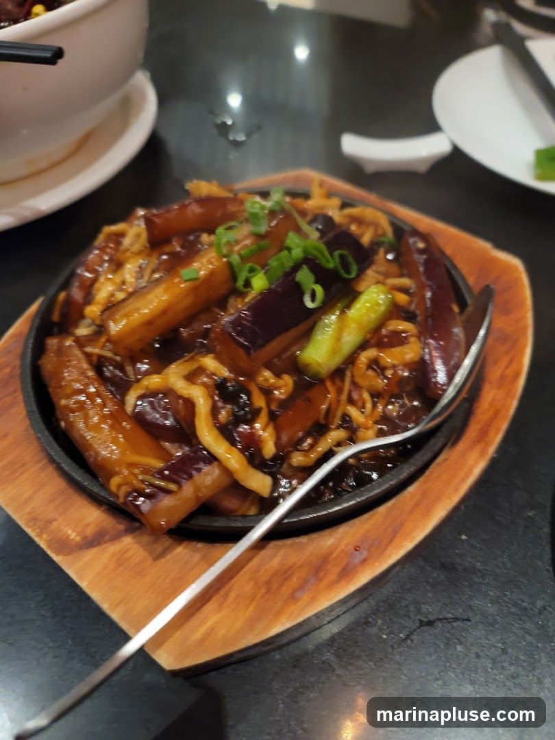 A close-up shot of a sizzling eggplant dish served on a black cast iron plate with a wooden base. The dish features pieces of eggplant and noodles coated in a dark, glossy sauce, garnished with chopped green onions. A fork rests on the side of the plate. In the background, a white bowl and a white plate are partially visible.