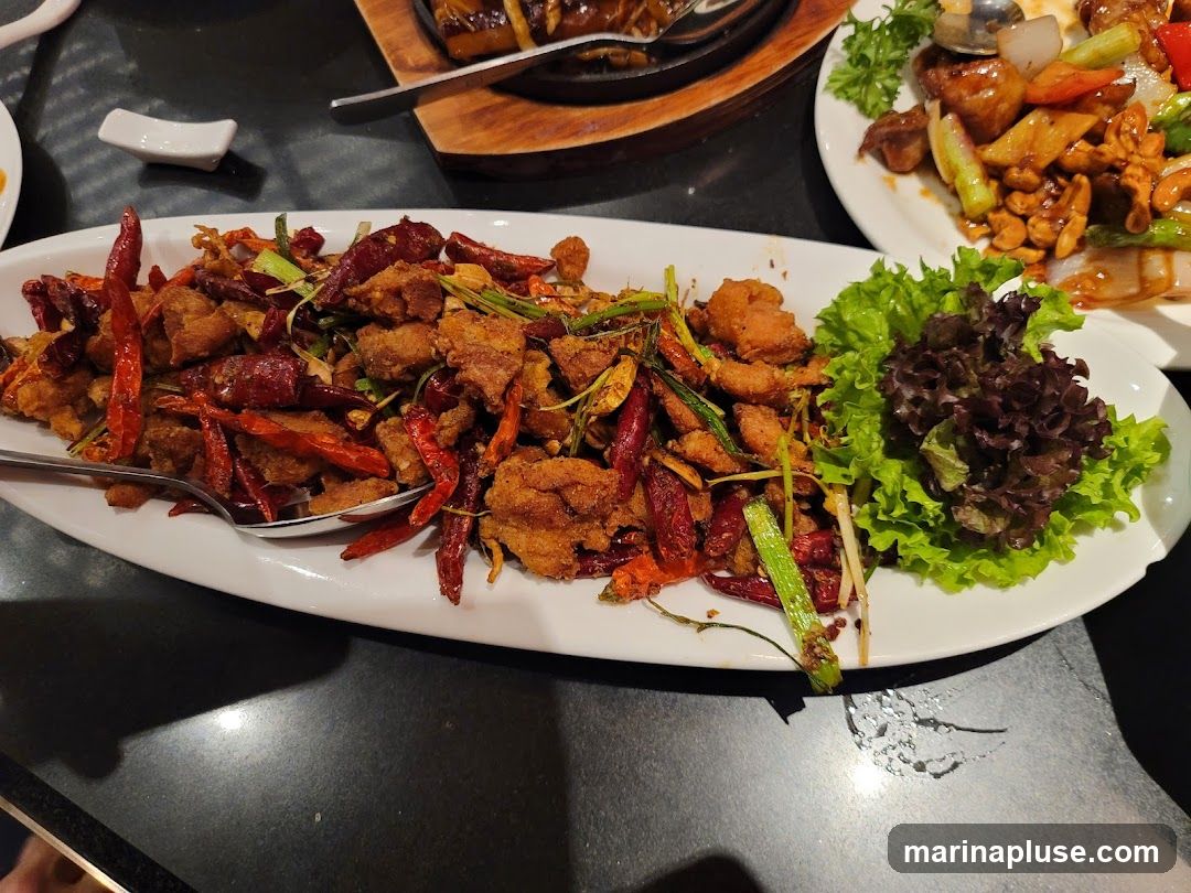 A platter of crispy fried chicken pieces tossed with dried red chilies and green onions, garnished with lettuce. Another dish with chicken, cashews, and vegetables is visible in the background.