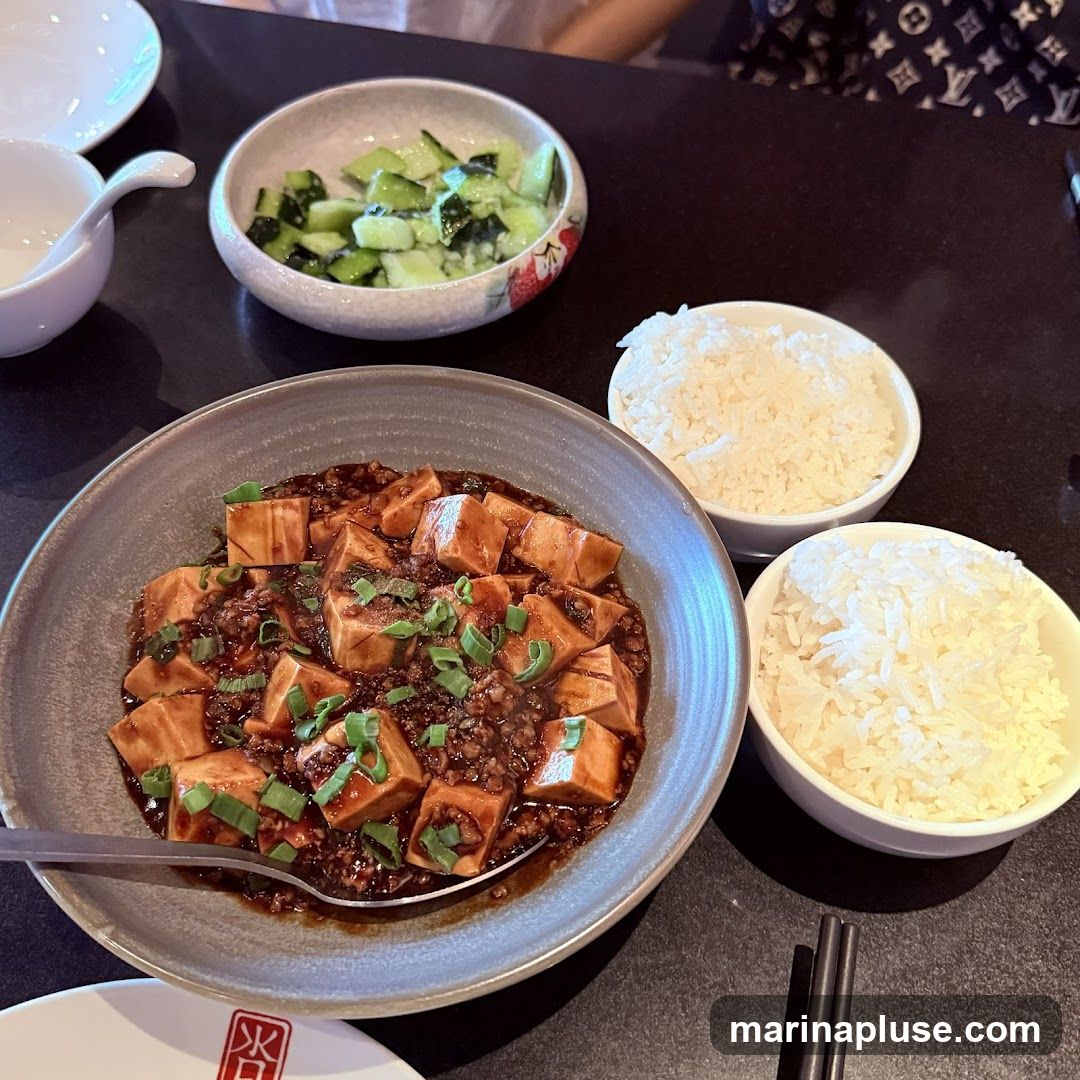 A close-up shot of a bowl of mapo tofu, a Sichuan dish made with tofu, minced meat, and chili bean paste, garnished with chopped green onions. Two bowls of white rice are placed next to the mapo tofu. A small bowl of sliced cucumbers is also visible.