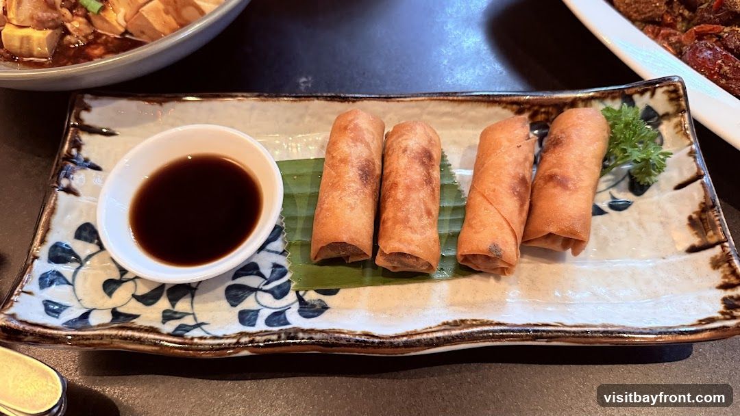 Four golden-brown spring rolls are arranged on a decorative rectangular plate with a small white dish of dark dipping sauce. A sprig of parsley garnishes the plate. In the background, another dish of food is partially visible.