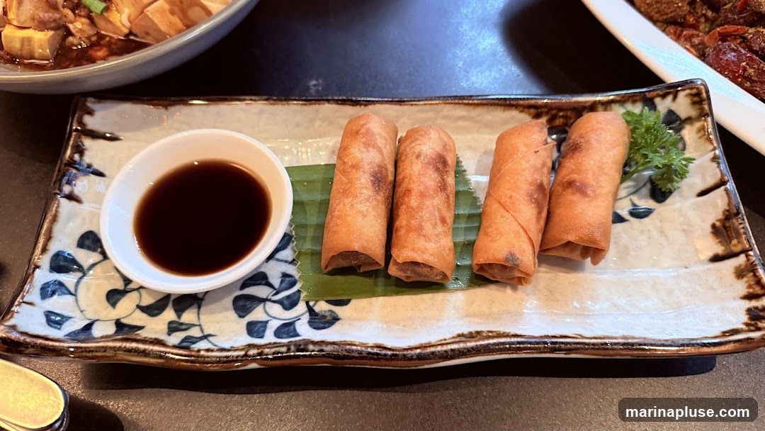 Four golden-brown spring rolls are arranged on a decorative rectangular plate with a small white dish of dark dipping sauce. A sprig of parsley garnishes the plate. In the background, another dish of food is partially visible.