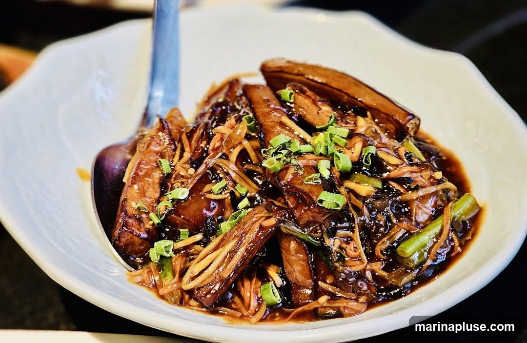 A close-up shot of braised eggplant served in a white bowl. The eggplant pieces are coated in a dark, glossy garlic sauce and garnished with chopped green onions. Thinly sliced vegetables, possibly bamboo shoots or mushrooms, are mixed in with the eggplant. A spoon is visible on the left side of the bowl.